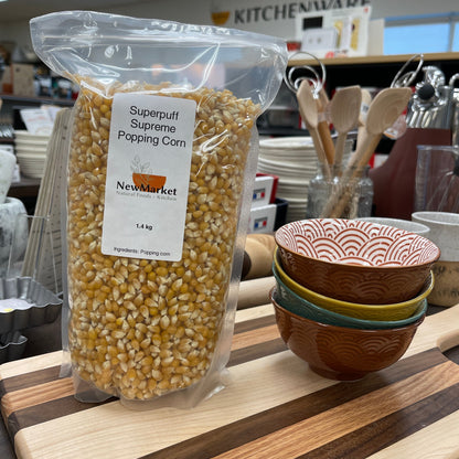 Bag of superpuff supreme popcorn kernels on a kitchen counter with a cutting board and kitchen utensils in the background.