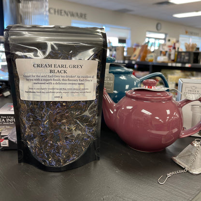 Package of cream earl grey black tea on a table with teapots and a diffuser.