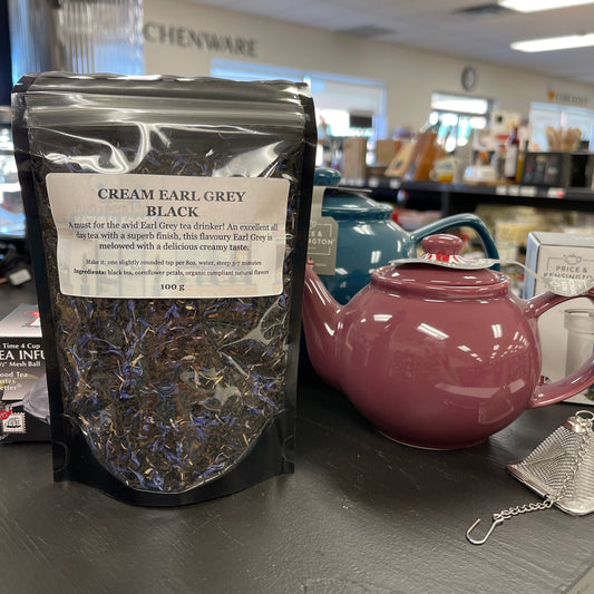 Package of cream earl grey black tea on a table with teapots and a diffuser.